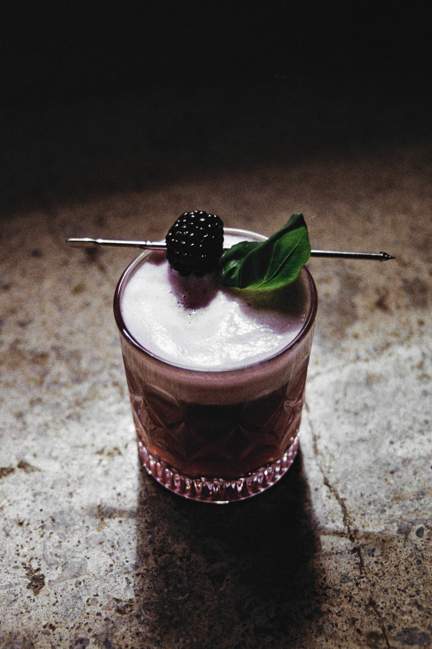 Image of the B&B Fizz cocktail, a blackberry and basil gin flip. Seen in a rocks glass the deep purple drink has a head of foam from the egg white.