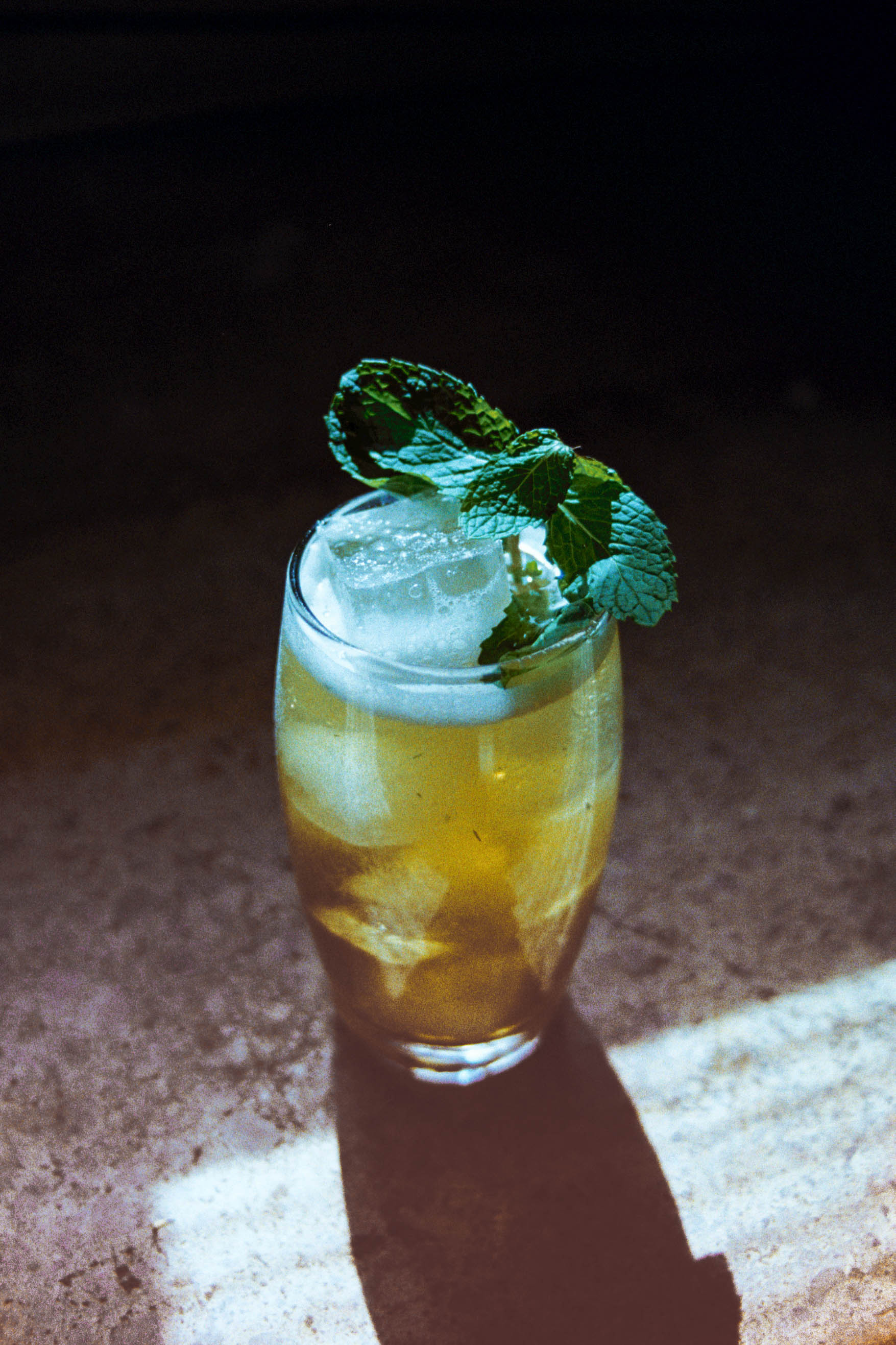 Boston Tea Derby cocktail in a highball glass filled with ice, garnish with mint on a wood table at a leather booth.