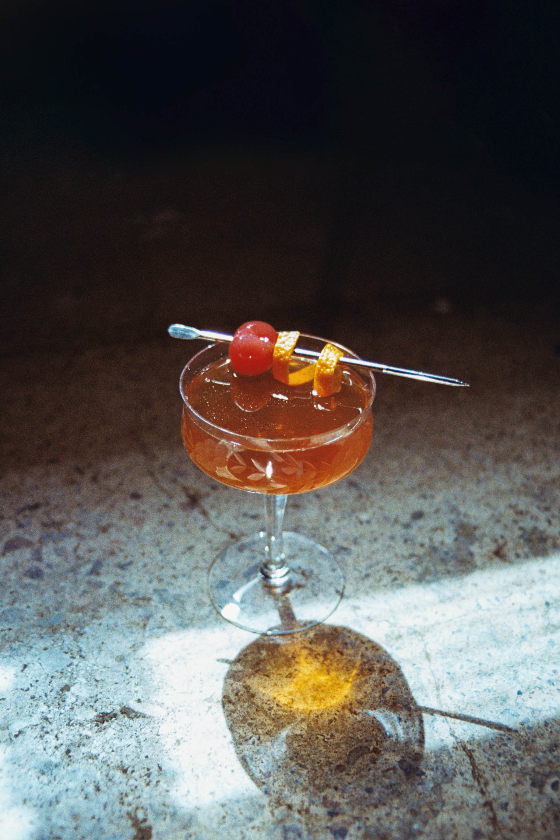 Image of the dark roast manhattan, a cocktail made using bourbon, coffee liqueur and bitters. Served up in a coupe with a cherry and orange twist.