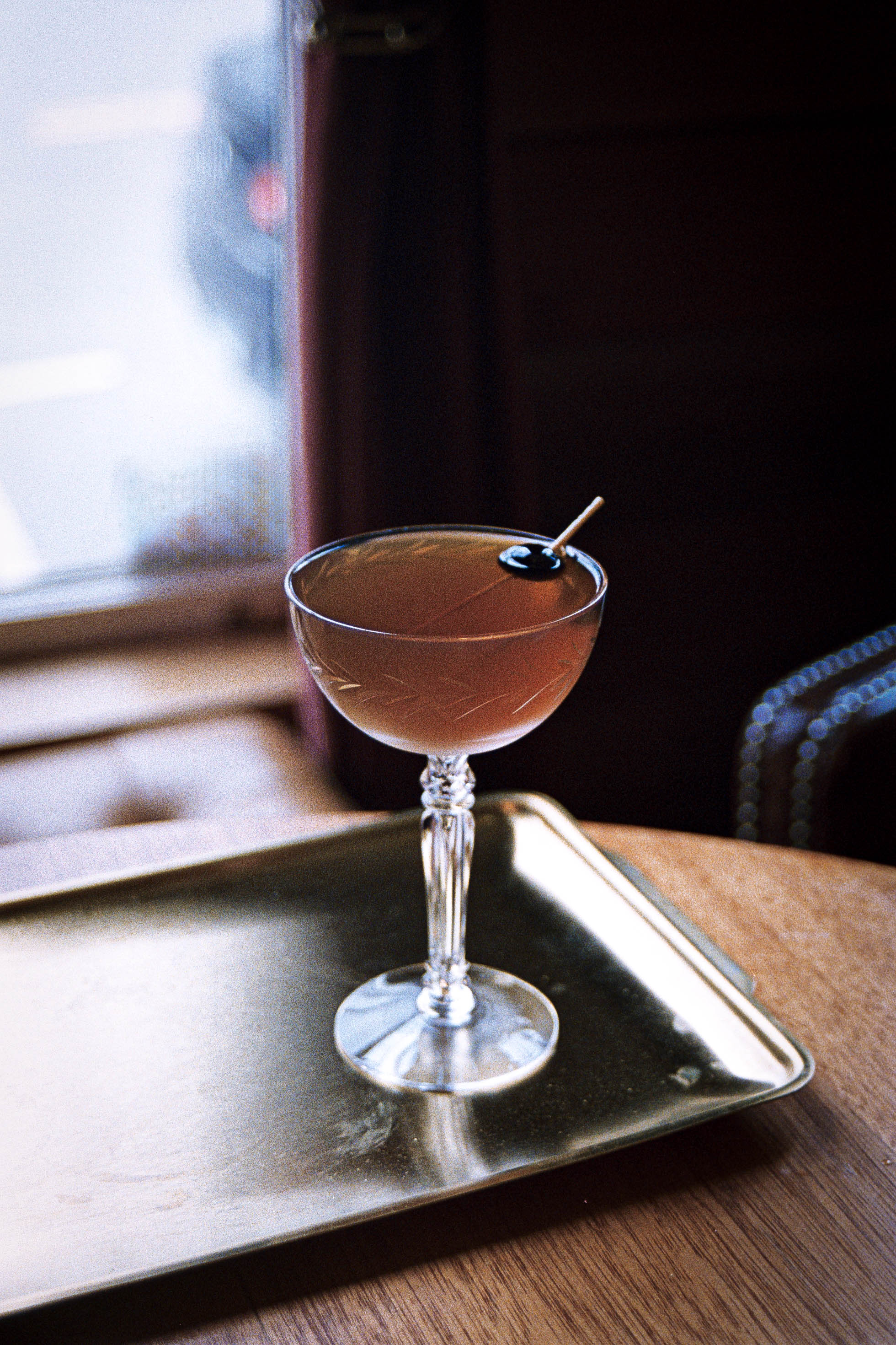 Close up shot of the Dark Roast Negroni cocktail in a vintage crystal coupe glass with a maraschino cherry garnish on a brass platter.